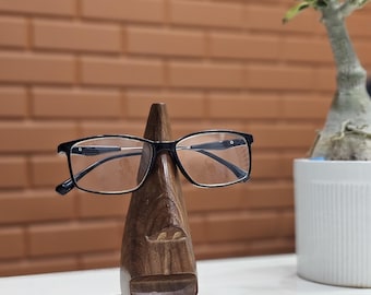 Puede incluir: Un soporte de gafas de madera con forma de rostro, con gafas de montura negra apoyadas. El soporte es de madera oscura y se asienta sobre una pequeña base cuadrada. El fondo incluye una pared de ladrillo y una planta en maceta.
