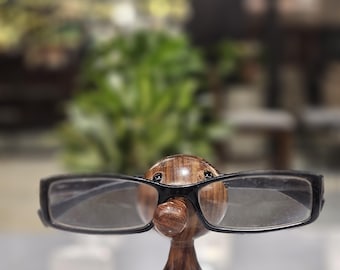 Puede incluir: Un soporte de gafas de madera con forma de pato, con un acabado marrón oscuro. El pato lleva unas gafas con montura negra. El soporte está sobre una superficie blanca, con un fondo borroso de vegetación.