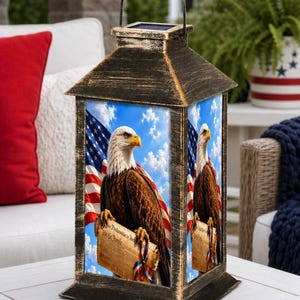May include: A rectangular solar lantern with a distressed black and gold finish, featuring a metal handle and a solar panel on top. The side panels display a bald eagle holding a scroll that reads We the People against an American flag background.