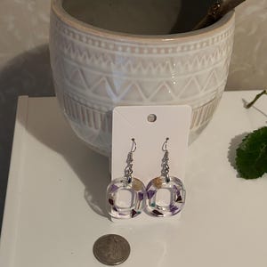 May include: Clear square resin earrings with silver hooks. The earrings contain small, colourful dried flowers and other decorative elements. A coin is next to the earrings for size comparison. A decorative ceramic pot is in the background.