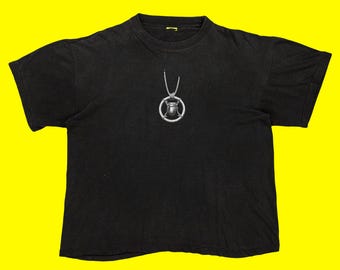 May include: Black t-shirt featuring a silver pendant with a beetle design. The pendant hangs from a thin chain. The t-shirt is a crew neck style and appears to be made of cotton or a similar fabric. The background is a bright yellow.