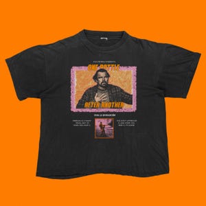 May include: Black t-shirt featuring a graphic from Paul Thomas Anderson's "One Bottle After Another." The design includes a portrait, text, and a small image. The shirt also has the phrases "Freedom is a funny thing, ain't it when you have it" and "You don't appreciate it and when you miss it, it's gone."