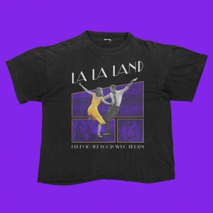 May include: Black t-shirt featuring the words "LA LA LAND" above a graphic of a dancing couple in front of a purple background. The phrase "HERE'S TO THE FOOLS WHO DREAM" is printed below the graphic.