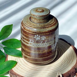 May include: A ceramic pet urn with a lid. The urn is cylindrical with brown and tan horizontal stripes. The word "Buddy" is engraved below a paw print design. The urn is sitting on a wooden slice.