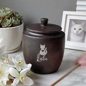 May include: A dark brown ceramic pet urn with a lid and a small knob. The urn features a cat silhouette with a heart and the name "Lulu" in script. A white framed photo of a white cat is in the background.