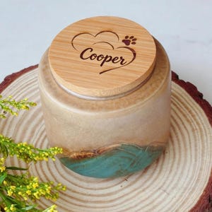 May include: A ceramic pet urn with a bamboo lid. The lid is engraved with the name "Cooper" inside a heart and paw print design. The urn has a tan and teal color scheme and sits on a wooden slice.