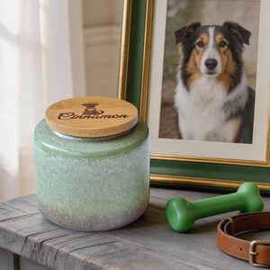 May include: A green ceramic pet treat jar with a bamboo lid, engraved with the word "Cinnamon" and a dog silhouette. A framed dog portrait, a green bone-shaped toy, and a leather dog collar are also visible.