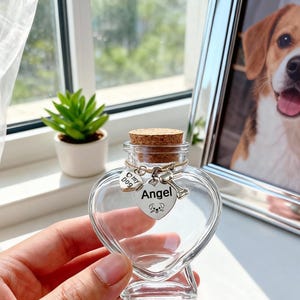 Op de afbeelding: Een heldere, hartvormige glazen fles met een kurken stopper. De fles heeft een zilveren bedel met de tekst "Angel" en "my dog". Een kleine groene plant in een witte pot en een ingelijst hondenportret staan op de achtergrond.