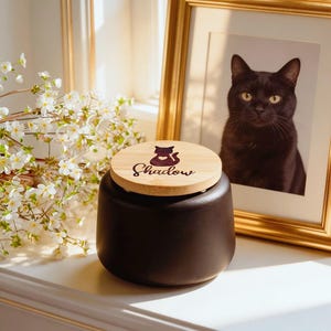 May include: A black ceramic pet urn with a wooden lid. The lid is engraved with a cat silhouette and the name "Shadow". A framed photo of a black cat is in the background.