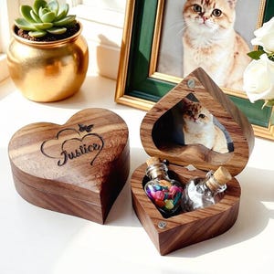 Könnte beinhalten: Zwei herzförmige Holzkisten. Eine geschlossene Kiste hat das Wort "Justice" auf dem Deckel eingraviert. Die andere Kiste ist offen und zeigt ein Foto einer Katze und zwei kleine Glasflaschen mit Korken. Eine Flasche enthält bunte Objekte.