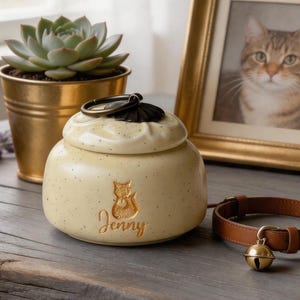 May include: A cream-colored pet urn with a cat silhouette and the name "Jenny" in gold script. The urn has a black handle and lid, and is next to a brown leather cat collar with a gold bell.