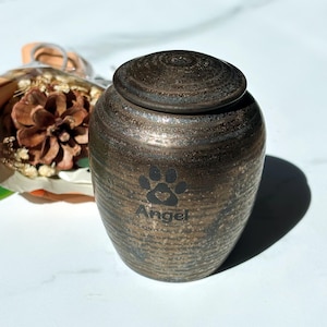 Könnte beinhalten: Bronze-farbene Urne für Haustiere mit Pfotenabdruck und dem eingravierten Namen "Angel". Die Urne hat einen Deckel und eine strukturierte Oberfläche. Eine dekorative Kiefernzapfen und Trockenblumen befinden sich im Hintergrund.