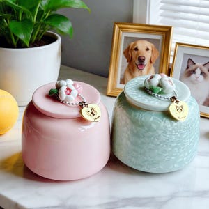 Op de afbeelding: Twee keramische potten met deksels, één in effen pastelroze en één in lichtgroen met een textuurafwerking. Elk deksel is voorzien van een wit bloemornament met groene bladeren en een gouden hartvormige bedel aan een kralenketting.