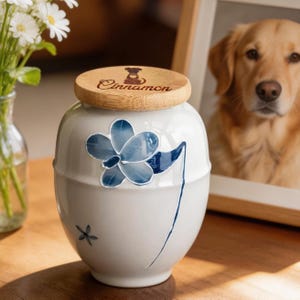 May include: A white ceramic pet urn with a wooden lid. The urn features a blue floral design and the word "Cinnamon". A framed photo of a golden retriever is in the background.