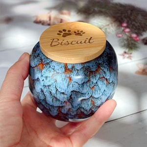 May include: A blue and orange ceramic jar with a wooden lid. The lid is engraved with paw prints and the word "Biscuit". The jar has a textured, glazed finish.