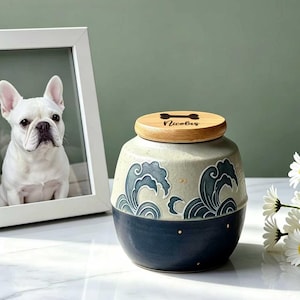 May include: A ceramic jar with a rounded shape, featuring a two-tone design with a cream-colored upper section decorated with dark blue wave patterns and a solid dark blue lower section. The wooden lid is engraved with a bone icon and the name Nicolas.