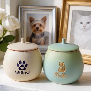 May include: Two ceramic pet urns, one cream-colored with a paw print and the name "Willow", the other green with a cat silhouette and the name "Muffin". Both have matching lids and are displayed near framed pet portraits.