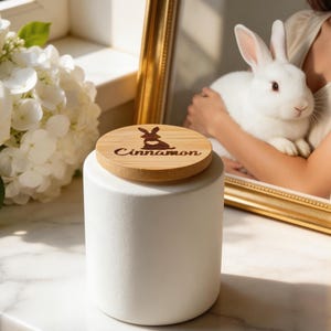 以下が含まれることがあります： 木製の蓋が付いた白いセラミックキャニスター。蓋にはウサギと「Cinnamon」の文字が刻まれています。キャニスターは円筒形で、大理石の表面に置かれています。背景には白いウサギの額縁写真があります。
