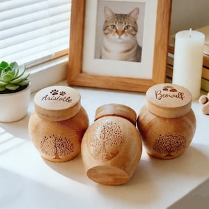 May include: Three wooden urns with engraved tree designs and wooden lids. The lids are engraved with the names "Aristotle" and "Beowulf". A framed cat portrait, a succulent plant, and a candle are in the background.