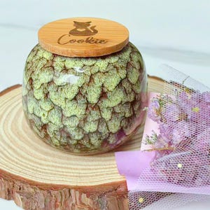 May include: A ceramic cookie jar with a wooden lid. The jar has a textured, green and brown pattern. The lid is engraved with a cat silhouette and the word "Cookie". The jar sits on a wooden slice.