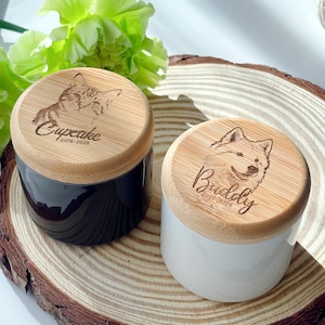May include: Two ceramic pet urns with wooden lids. One urn is black, the other is white. Each lid features an engraved pet portrait and name, with the years of life. The urns are displayed on a wooden slice.
