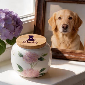 Puede incluir: Una urna para mascotas de cerámica blanca con tapa de madera con silueta de perro y el nombre "Buddy". La urna está decorada con flores de hortensia rosas y hojas verdes. En el fondo, una foto enmarcada de un golden retriever.