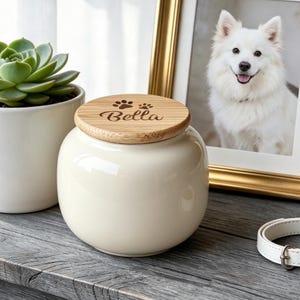 May include: A cream-coloured ceramic jar with a round bamboo lid. The lid features the engraved name Bella and two paw prints. The jar has a smooth, glossy finish and a rounded shape, serving as a container for small items or keepsakes.