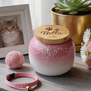 May include: A pink and white pet urn with a wooden lid engraved with the name "Bella" and paw prints. A pink cat collar with a gold bell, a ball of pink yarn, and a framed cat portrait are also visible.