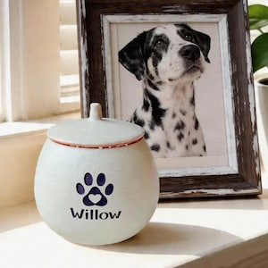May include: A cream-coloured pet urn with a paw print and heart design, the name "Willow" is printed below. The urn has a lid with a red rim. A framed black and white photo of a dog is in the background.