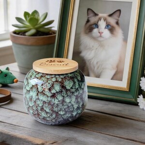 May include: A round ceramic jar with a textured, mottled green and brown glaze. The jar features a circular light-coloured wooden lid engraved with two small paw prints and the name "Biscuit" in a cursive script font on the top surface.