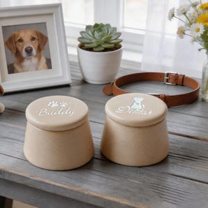May include: Two beige pet urns with lids, one labeled "Buddy" with paw prints and the other "Delilah" with a dog silhouette. A framed dog portrait, a succulent, and a brown leather collar are also visible.