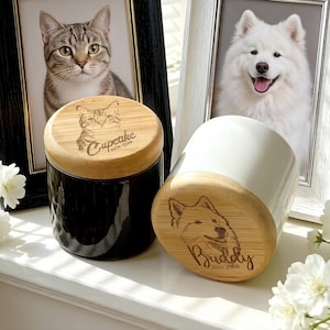 Peut inclure: Deux urnes pour animaux de compagnie avec des couvercles en bambou, une noire et une blanche. L'urne noire porte un portrait de chat avec le nom "Cupcake" et des dates. L'urne blanche porte un portrait de chien avec le nom "Buddy" et des dates. Portraits d'animaux encadrés en arrière-plan.