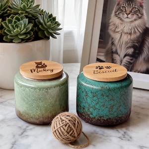 May include: Two cylindrical ceramic jars with bamboo lids. The left jar is light green with a brown base and a cat icon with the name Mickey. The right jar is teal with a dark crackle pattern, a brown base, and paw prints with the name Biscuit.