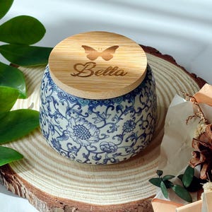 May include: A ceramic jar with a blue and white floral design, topped with a wooden lid. The lid features a butterfly design and the name "Bella" engraved. The jar sits on a wooden slice.