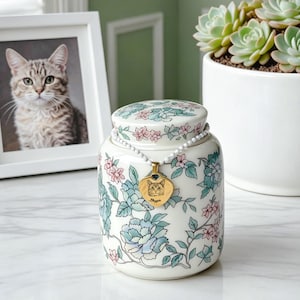 May include: A cylindrical ceramic urn with a white base featuring a floral pattern in shades of blue, green, and pink. A white beaded necklace with a gold heart-shaped pendant engraved with a cat illustration and the name Maggie rests on top.