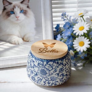 May include: A ceramic jar with a wooden lid, engraved with a butterfly and the name "Bella". The jar displays a blue and white floral design. A fluffy white cat with blue eyes is in the background, alongside a bouquet of daisies and blue flowers.
