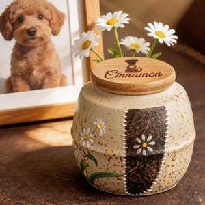 May include: A beige ceramic pet urn with a wooden lid, featuring the word "Cinnamon" and a dog silhouette. The urn is decorated with daisy and leaf designs, and a patch-like design. A framed photo of a dog is in the background.