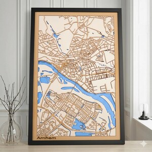 Op de afbeelding: Een houten kaart van Arnhem, Nederland, met een zwarte lijst. De kaart heeft een gedetailleerd lasergesneden ontwerp van de straten en waterwegen van de stad, met blauw dat het water voorstelt. Het woord "Arnhem" staat onderaan.