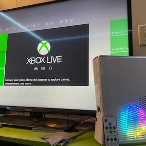 Könnte beinhalten: Eine weiße Xbox 360-Konsole mit Fernbedienung, neben einem Philips-Fernseher, der die Xbox Live-Oberfläche anzeigt. Die Konsole hat eine farbenfrohe Lichtanzeige. Der Fernseher zeigt den Text "XBOX LIVE" und Optionen zur Internetverbindung.