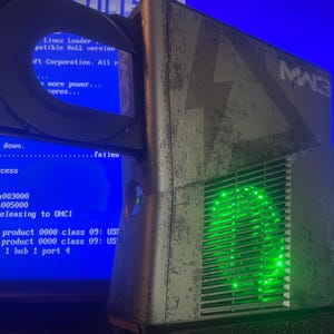 Könnte beinhalten: Eine graue und schwarze Spielkonsole mit abgenutztem Finish und dem Schriftzug MW3 an der Seite. Die Konsole verfügt über eine grün beleuchtete kreisförmige Lüftung und ein offenes Laufwerk. Ein blauer Bildschirm mit weißem Text ist zu sehen.