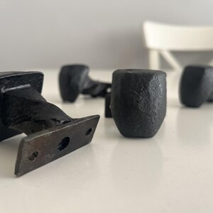 May include: Four black cast iron cabinet pulls with a textured finish. Each pull has a square head and a rectangular base with pre-drilled holes for mounting. These vintage-style pulls are designed for easy installation and add a rustic touch.