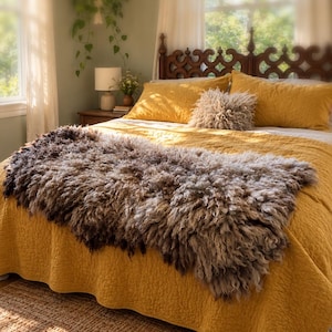 May include: A mustard yellow quilted bedspread covers a bed, topped with a long, shaggy, grey and brown sheepskin throw. Two matching mustard yellow pillows and a small, textured, shaggy grey accent pillow are arranged against a dark wood headboard.