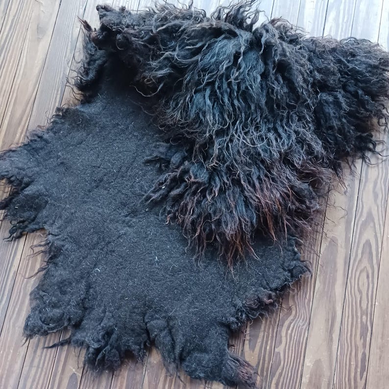 Puede incluir: Una alfombra de piel de oveja gris oscuro con un aspecto peludo y texturizado. La alfombra tiene una forma natural e irregular y una mezcla de fibras cortas y largas. El color pasa del gris oscuro a un marr&oacute;n m&aacute;s claro en algunas zonas.