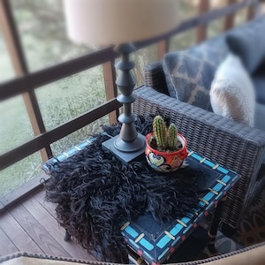 May include: A small, black table with a decorative border, topped with a black, shaggy throw. A gray lamp and a colorful, patterned pot with cacti sit on the table. A wicker chair and outdoor setting are in the background.