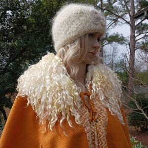May include: A handcrafted outfit comprising a cream-coloured, fuzzy hat with a floral detail, and a rust-coloured cape. The cape has a decorative cream trim and a fluffy, cream-coloured collar.