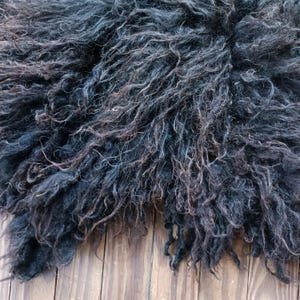 Puede incluir: Primer plano de una alfombra de piel de oveja oscura con fibras largas y rizadas. La alfombra presenta tonos negros y marrones, creando una textura densa y suave. Se encuentra sobre una superficie de madera.