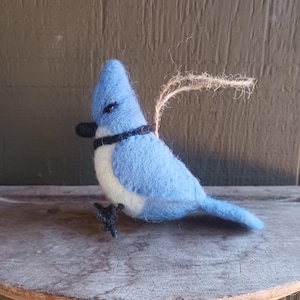 Puede incluir: Un adorno de pájaro de fieltro azul con vientre blanco y ojos negros. El pájaro tiene una banda negra alrededor del cuello y un colgador de cuerda marrón.