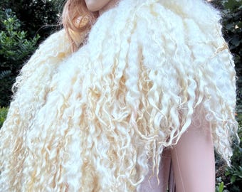 May include: A cream-colored, long-haired, curly textured shawl draped over the shoulders. The garment features dense, flowing locks of wool-like fiber that cascade down the front, creating a voluminous and layered appearance.