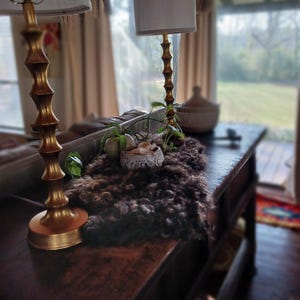 May include: A dark brown, shaggy textile runner adorns a dark wooden console table. Two gold-toned lamps with white shades flank the runner. A small decorative bowl and greenery are placed on the runner, creating a cozy, rustic aesthetic.