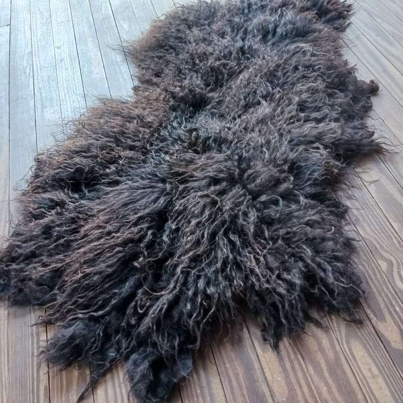Puede incluir: Una alfombra de piel de oveja gris oscuro con fibras de lana largas y rizadas. La alfombra tiene una forma irregular y est&aacute; sobre un suelo de madera. El color var&iacute;a de gris oscuro a marr&oacute;n, creando una apariencia natural y texturizada. Para la decoraci&oacute;n del hogar.